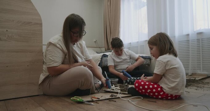 Caring mother with her son and daughter sitting on the floor, creating a diy project together with a hot glue gun, enjoying a creative family activity and quality time in the living room.