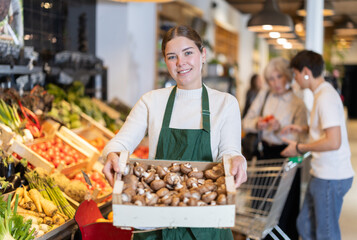 Obraz premium Girl employee in dark green apron stands in sales area with box of mushrooms. Employee carries box of mushrooms from storage area into sales area.