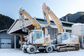 Scenic view frozen hydraulic excavators standing outdoors winter conditions covered snow ice industrial building alpine area. Cold weather impacts machinery operation hydraulic systems reliability