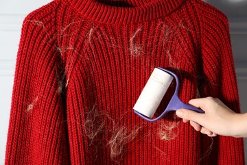 Woman cleaning sweater with lint roller near light grey wall indoors, closeup