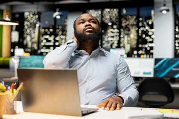 Black male professional does self massage to relieve neck stiffness and discomfort, dealing with tension due to incorrect office posture after hours. Stretching his upper back muscles.
