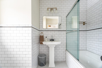 Bright white bathroom interior with subway tile walls, pedestal sink and glass shower enclosure,...