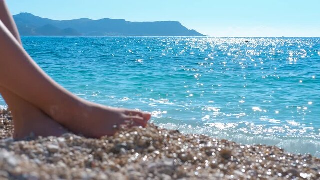 Woman relaxing on pebble beach with sparkling turquoise sea. Female legs and bare feet relaxing on a sunny pebble beach with turquoise water lapping the shore, a perfect summer vacation
