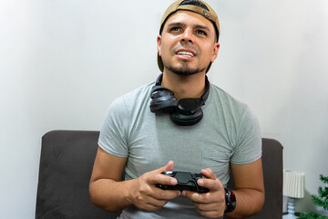 Man sitting on couch focused on video game while holding controller, wearing headphones and backward cap