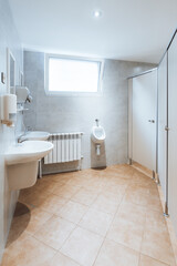 Wide-angle view of clean modern public restroom with tiled floor, wall-mounted sink, urinal, toilet stall partition, soap dispenser and bright window, minimal interior with copy space