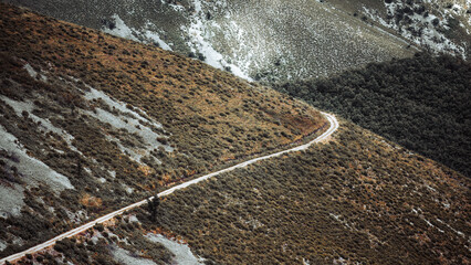 Winding dirt road crossing rugged hillside with scrub vegetation and rocky patches, aerial landscape view of remote mountain terrain, natural texture background and copy space