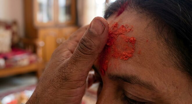 Elder's hand applying a vibrant red pigment tilak to a youth's forehead