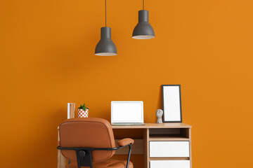 Workplace with blank laptop and lamps near orange wall