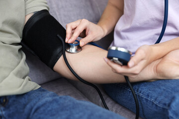 Woman checking man's blood pressure with sphygmomanometer on sofa at home, closeup