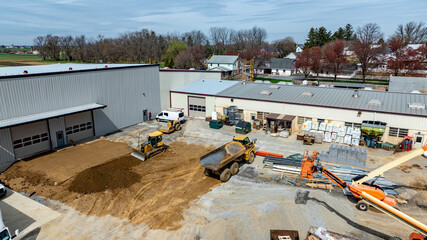 Construction equipment is busy at a site with trucks, loaders, and piles of materials. Workers are moving dirt and supplies in an area near buildings and trees under a clear sky.