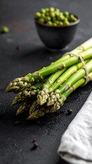 Bundled asparagus spears sit near a bowl of green peas against a dark textured surface with a light fabric