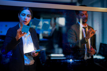 Diverse corporate team brainstorming ideas on glass wall. Businessman writing project plan with marker while female colleague analyzes sticky notes in modern dark office at night.