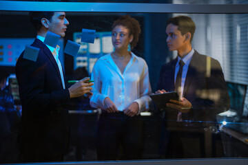Diverse corporate team brainstorming ideas on glass wall. Businessman writing project plan with marker while female colleague analyzes sticky notes in modern dark office at night.