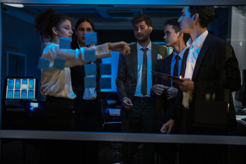 Agile tech startup team conducting scrum meeting on glass wall. Developers and managers mapping software workflow with sticky notes in modern blue-lit office at night.