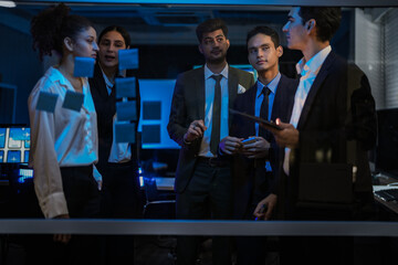 Agile tech startup team conducting scrum meeting on glass wall. Developers and managers mapping software workflow with sticky notes in modern blue-lit office at night.