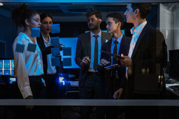 Agile tech startup team conducting scrum meeting on glass wall. Developers and managers mapping software workflow with sticky notes in modern blue-lit office at night.