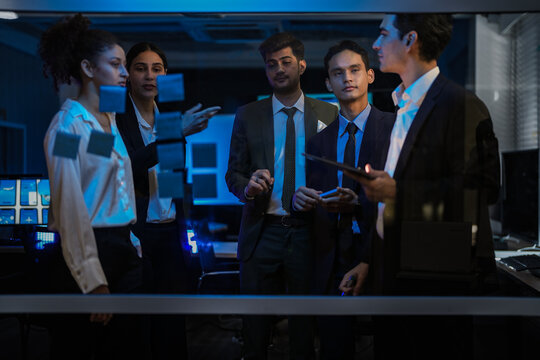 Agile tech startup team conducting scrum meeting on glass wall. Developers and managers mapping software workflow with sticky notes in modern blue-lit office at night.