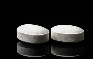 Two white oval tablets on a glossy black surface with strong reflections, minimalist clinical composition conveying a calm sterile mood