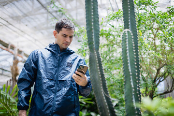 Young scientist in farm greenhouse uses smartphone for agricultural research, focusing on cactus plants as part of dedicated research team working in farm environment