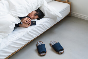 布団にくるまりスマートフォンを操作している若い男性　ライフスタイルイメージ
