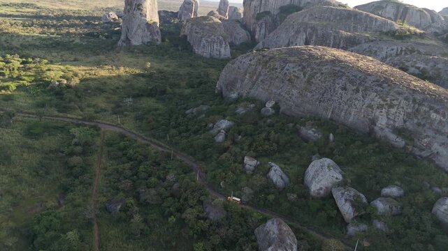 Low-angle cinematic drone flyby of the ancient black rocks at Pungo Andongo, Angola, yellow camper van truck ovelanding Africa travelling in narrowed road between rock formation panoramic aerial