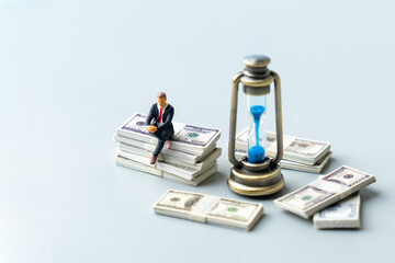 Businessman sits on a stack of cash bills next to an hourglass. Financial planning, retirement, savings, return on investment concept.