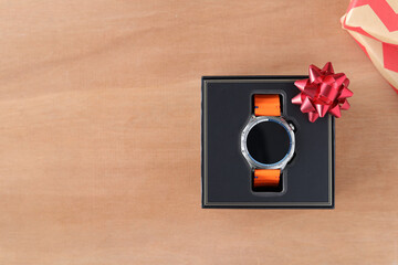 Top view of a smartwatch with an orange band in a premium black gift box, with a red bow on wooden background. Flat lay with copy space for promotion and christmas, new year gift.