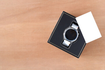 Top view of a modern smartwatch in a luxury black gift box. A blank white card , ready for a message on a wooden background with copy space. Perfect for holidays or celebrations.