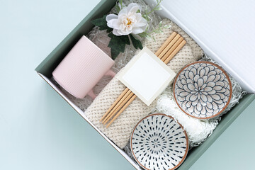 Concept of housewarming, or thank-you present. Top view gift box featuring a cozy knitted blanket, a pink ceramic mug, chopsticks, and two ceramic bowls with unique floral patterns on blue background