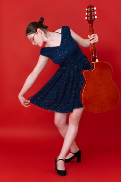 Female guitarist signs off with curtsy and bow, thanking audience for applause with eyes closed. Musician attired in blue above knee dress and high heels. 18 year old singer songwriter wearing glasses