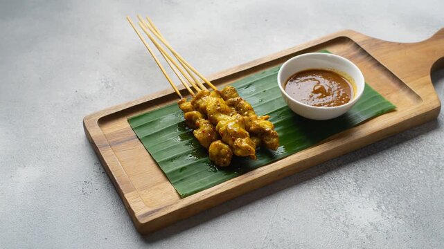 Grilled chicken satay with peanut sauce on a banana leaf and wooden board, slow 360-degree orbit shot, popular Southeast Asian appetizer 4k footage.