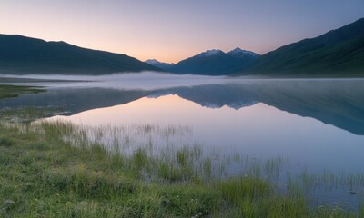 Obraz premium Calm lake at dawn reflects mountains under a soft, pastel sky; gentle mist