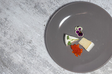 Minimalist gourmet plate with cheese, fish roe, and edible flower garnish on gray surface