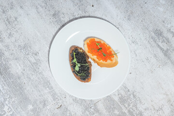 Two types of caviar on toasted bread garnished with herbs on a white plate, placed on a textured surface.