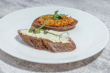 Two slices of rye bread topped with mustard and cream cheese on a white plate, garnished with greens on a textured background.
