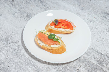 Two gourmet bruschetta appetizers on a white plate with toppings of smoked salmon, cream cheese, and fresh tomato, garnished with herbs on a textured background.