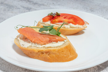 Close-up of two gourmet bruschetta slices topped with cream cheese, smoked salmon, tomato, and fresh herbs on a white plate, served on a textured background.