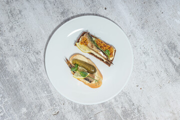 Top view of gourmet appetizers with fish, pickles, and mustard on a white plate against a rustic background.