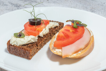 Two assorted open-faced sandwiches on a white plate, one with dark bread, cream cheese, tomato, and olive, the other with white bread, cream cheese, ham, and tomato.