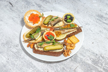 An appetizing plate of assorted canap&eacute;s with fish, pickles, nuts, and crackers presented on a textured background.