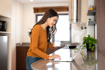 Smiling happy woman drinking coffee and looking at  laptop computer standing at kitchen table, relaxing at casual home.