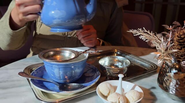 Close up of female hands pouring hot herbal tea from elegant blue ceramic teapot through silver strainer into matching cup on marble table in cozy cafe atmosphere