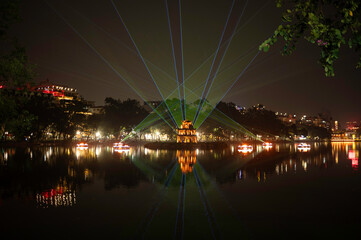 Naklejka premium Vietnam, Hanoi - Turtle Tower illuminated at night on Hoàn Kiếm Lake in Hanoi during a light show, with city buildings visible in the background