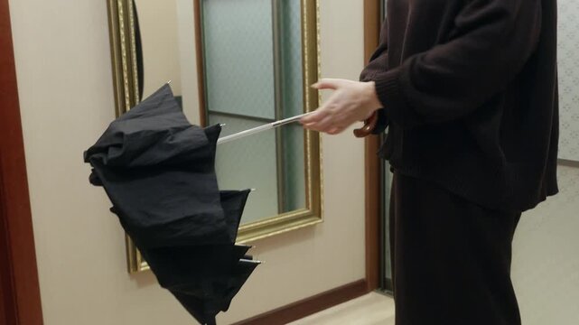 Young woman in dark sweater opening black umbrella indoors near mirror, practical calm mood symbolizing weather preparation and daily routine.