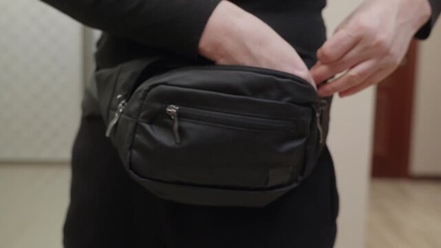 Close up of young woman zipping and unzipping black fanny pack indoors, focused casual mood symbolizing everyday organization and modern lifestyle.