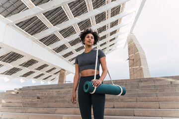 Active young woman with yoga mat ascending stairs beneath urban solar panel roof