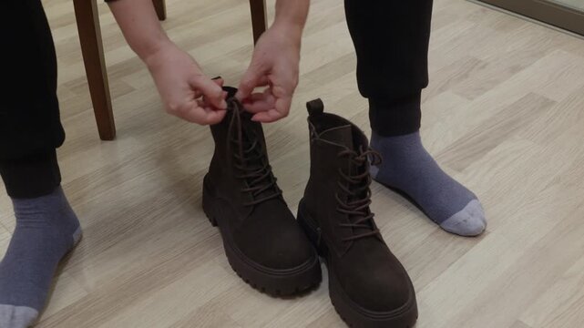 Young woman sitting on chair at wooden floor untangling brown shoelaces on boots indoors, focused calm mood symbolizing preparation and everyday routine.