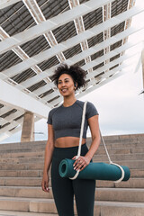 Fitness instructor walking with yoga mat under modern solar panel canopy, wearing wireless earbuds and ready to teach a yoga class