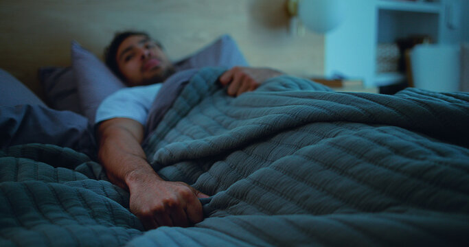 Insomnia, thinking and man in bed at night for burnout, exhausted and fatigue for mental health. Blanket, home and person with thoughtful, anxiety and reflection for sleepless, bad dream and tired