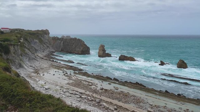 Slow-motion drone footage over the rocky coast of Arn&iacute;a, Cantabria, showing straight stratigraphic layers on the ground, eroded rocks, turquoise waves crashing over stones under a cloudy blue sky.